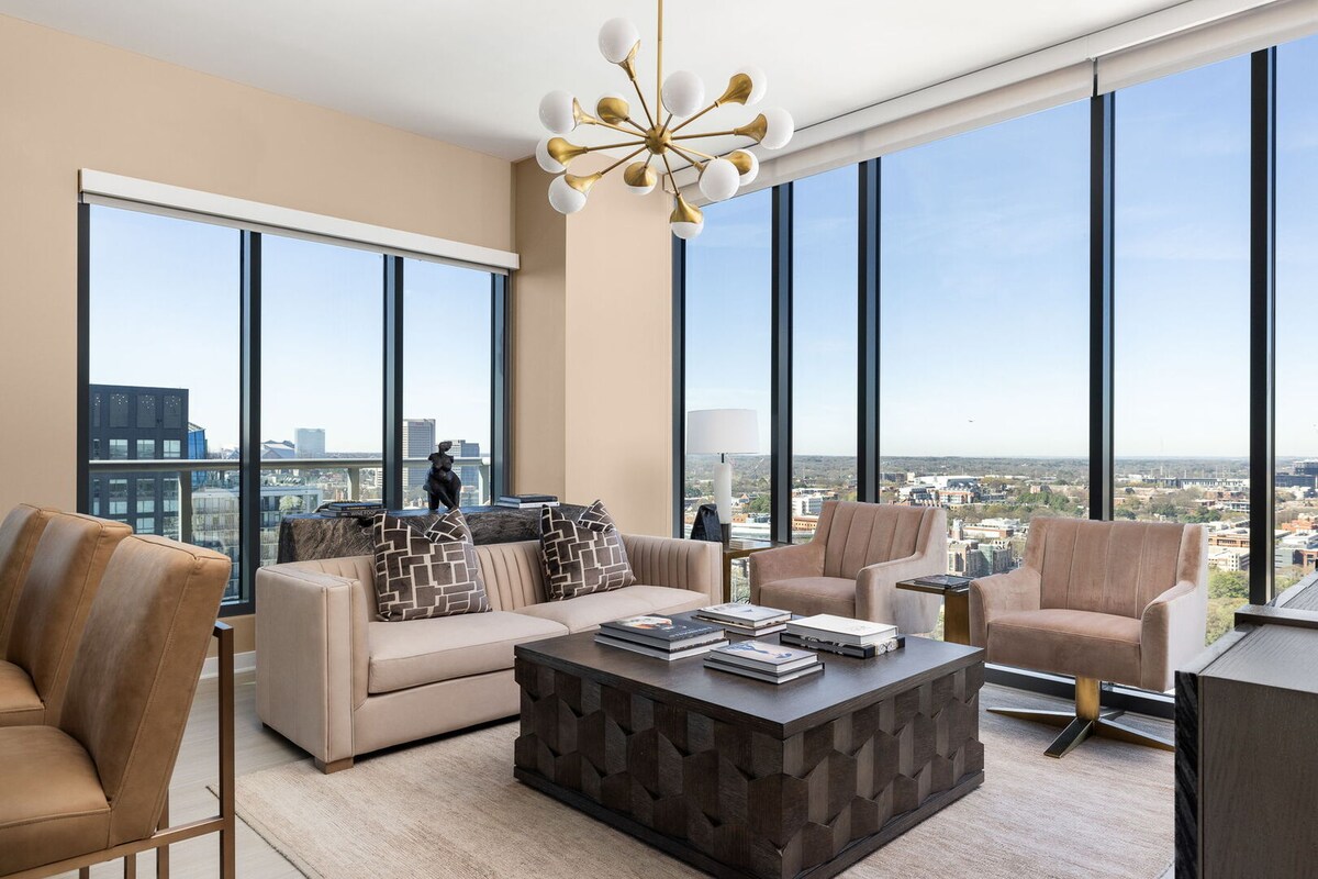 A modern living area showcases a large, plush sofa accompanied by two chic armchairs. A sleek coffee table sits centrally, adorned with decorative books. Floor-to-ceiling windows provide expansive views of the city skyline, flooding the space with natural light.