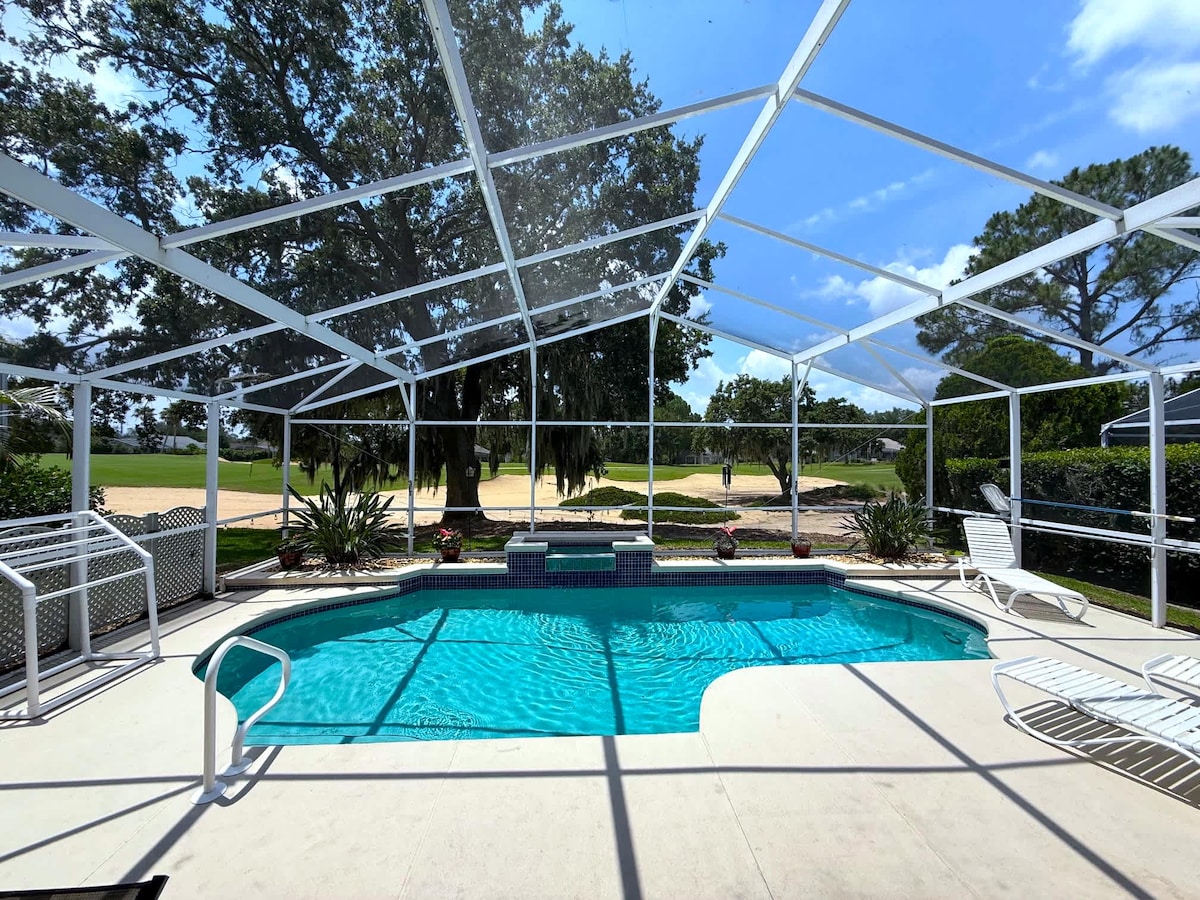The private pool is enclosed by a screened lanai, featuring a generous seating area around the water. Lush greenery and manicured landscaping frame the pool, while a golf course vista is visible beyond the enclosure. Bright blue skies and scattered clouds enhance the sunny ambiance.