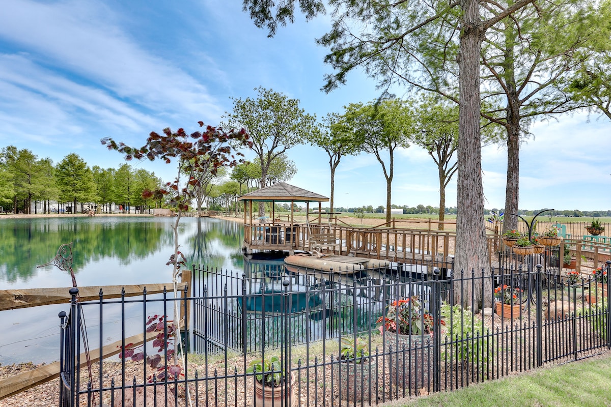 A waterfront gazebo is situated on the edge of a serene pond, surrounded by vibrant trees and greenery. A fence encloses the area, while a wooden deck extends toward the water, providing a peaceful outdoor retreat.