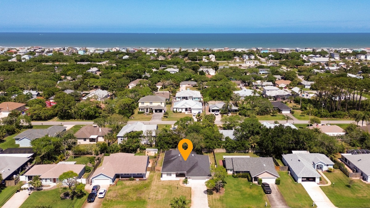 An aerial view captures a tranquil coastal neighborhood surrounded by lush greenery. The sandy beach and the ocean are visible in the distance, contrasting with the residential properties below. A highlighted location marker indicates the property from which the image is taken.