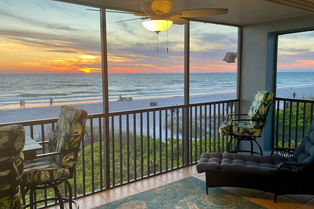A furnished balcony offers comfortable seating and a table, with expansive views of the beach and sunset. The horizon displays vibrant colors as the sun sets over the Gulf of Mexico, while the gentle waves wash onto the shore. Lush greenery is visible below.