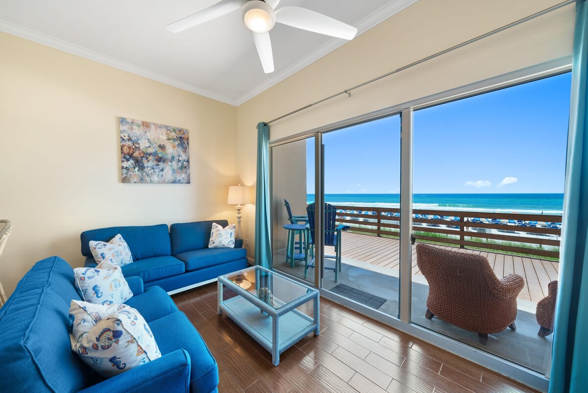 A welcoming living area features comfortable blue seating, including a sofa and accent chair, arranged around a glass coffee table. Large sliding glass doors provide a clear view of the outdoor patio and the Gulf beyond, inviting natural light into the space.