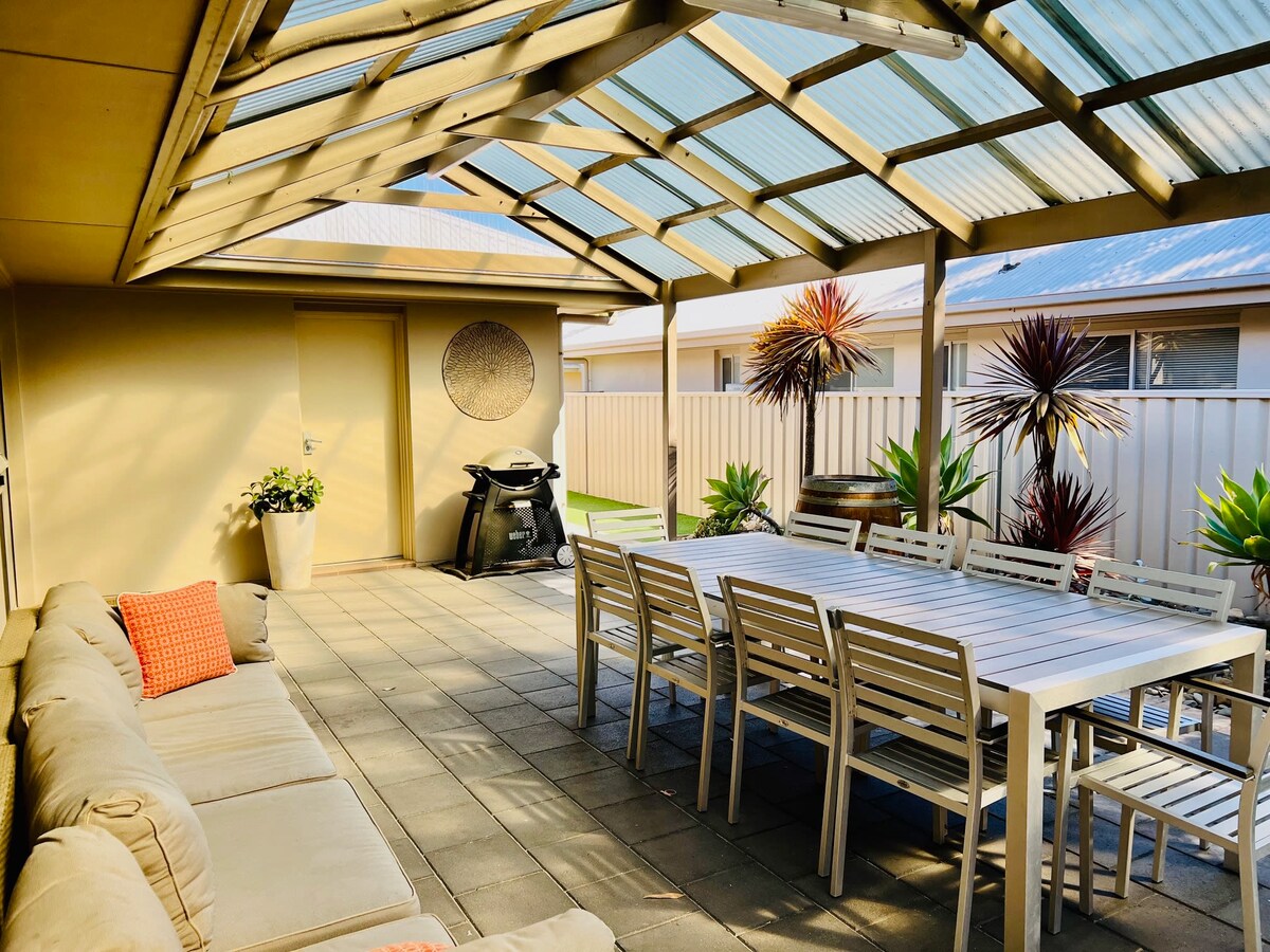 An outdoor pergola area is shown, featuring a large dining table surrounded by multiple chairs. A comfortable lounge area is visible to one side, along with a barbecue grill positioned nearby. Potted plants add greenery to the space, enhancing its welcoming appearance.