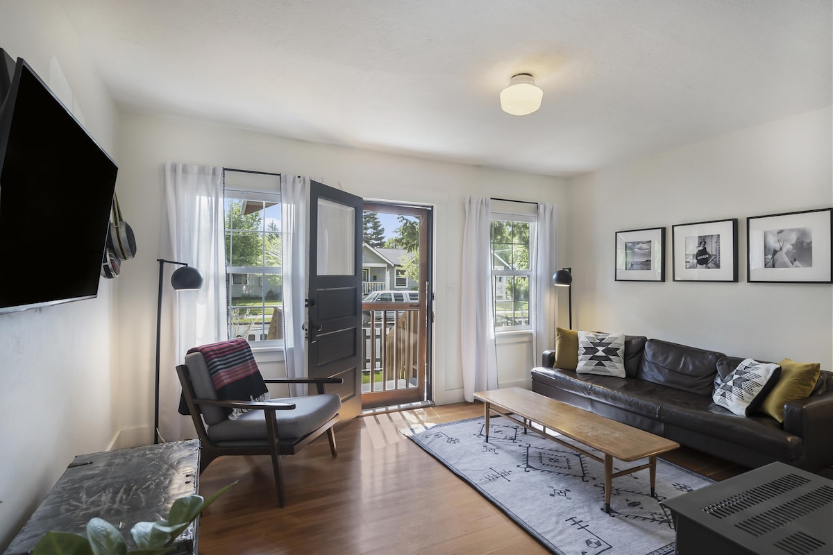 A light-filled living space features large windows framed by sheer curtains, allowing natural light to brighten the room. A comfortable leather sofa is positioned across from a television mounted on the wall. A wooden coffee table adds warmth, complemented by a stylish chair nearby.