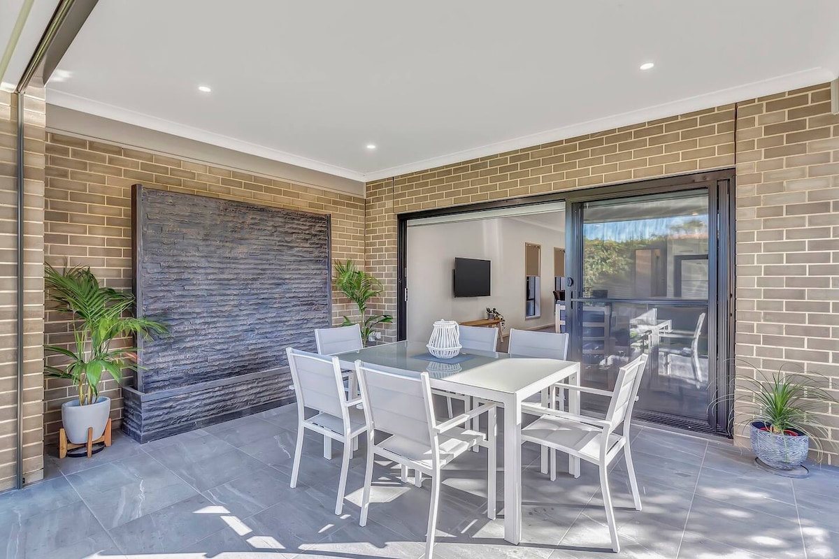 An outdoor dining area is presented with a sleek white table surrounded by matching chairs. A feature wall with textured tiles adds depth, while potted plants bring a touch of greenery. Large sliding doors provide access to the indoor space, enhancing light and openness.