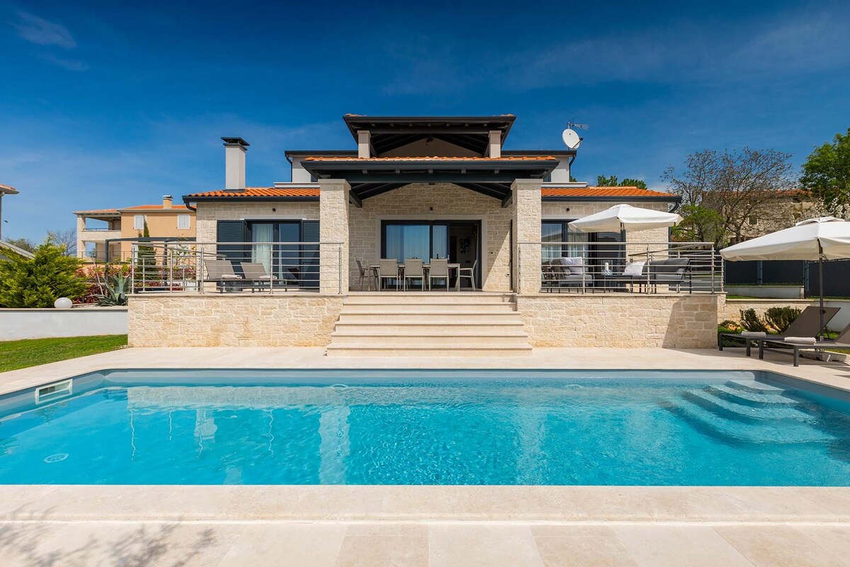 A modern villa is presented with a clean stone façade and spacious outdoor patio. A private pool glistens in the foreground, complemented by sun loungers. The entrance features wide steps leading to a covered terrace area, ideal for relaxation and outdoor dining.