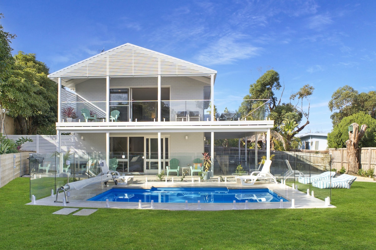 The exterior view showcases a two-story home surrounded by lush greenery. A clear pool is visible in the yard, accompanied by sun loungers and a spacious deck. The upper balcony features outdoor seating, providing a relaxing space overlooking the backyard and pool.