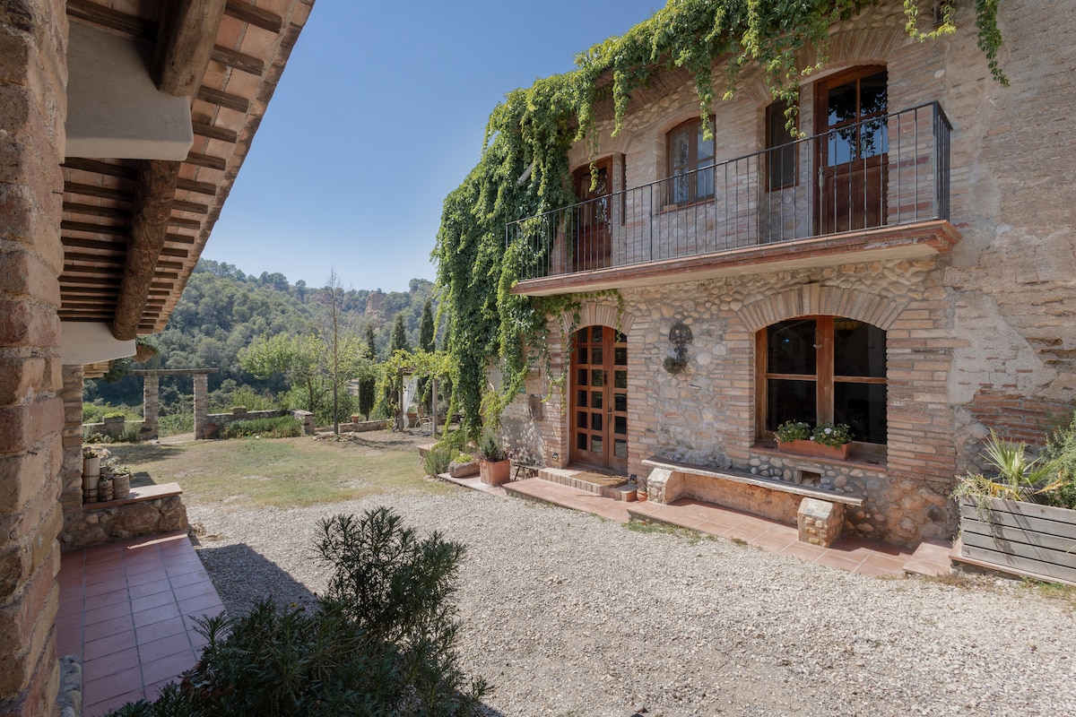 A charming farmhouse exterior is framed by lush greenery, showcasing stone walls and wooden accents. An inviting patio area is visible, featuring terracotta tiles and a seating bench. The surrounding landscape includes trees and a well-maintained garden, enhancing the serene atmosphere.