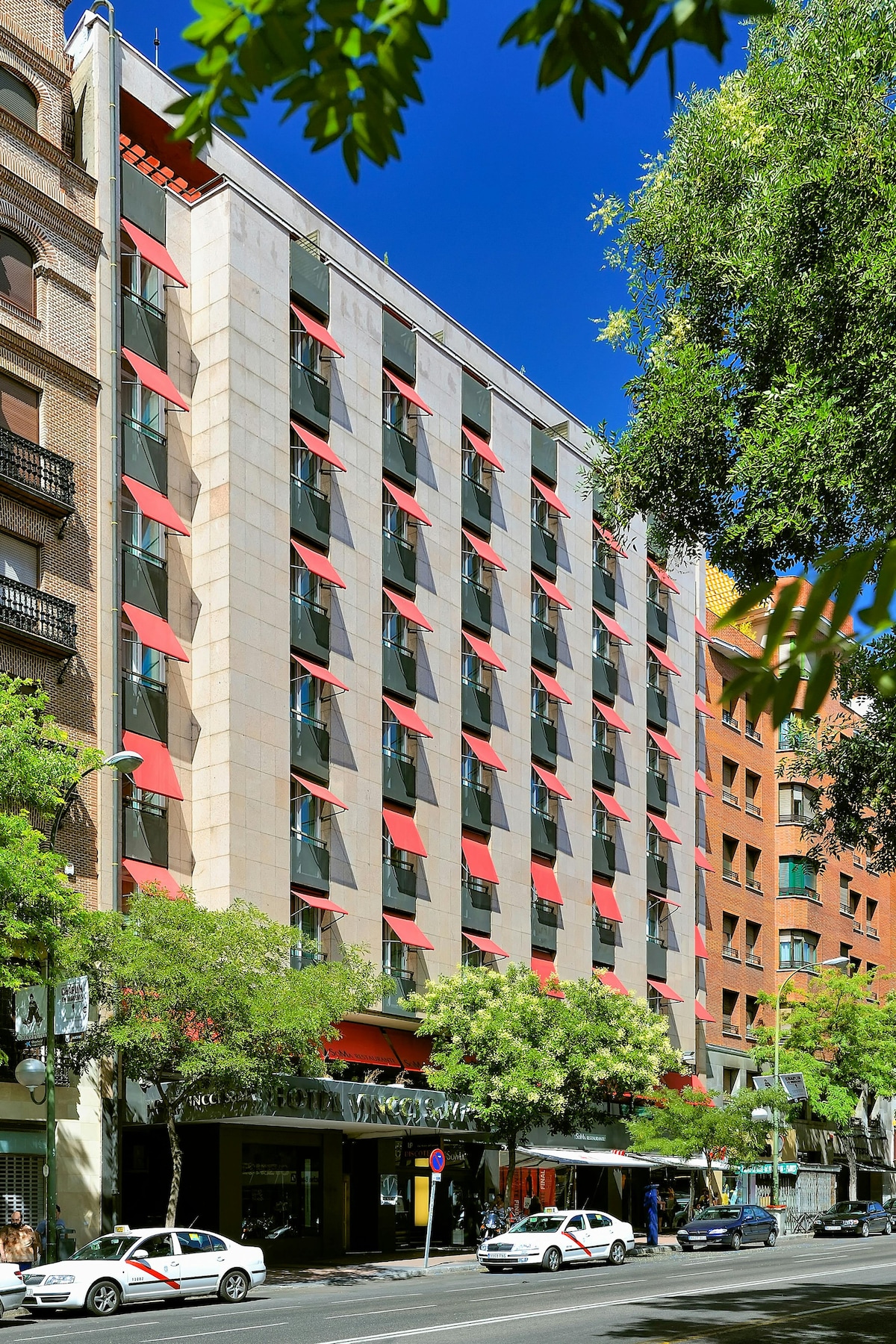 The hotel exterior showcases a modern façade with large windows and vibrant red awnings. Surrounding greenery and urban elements contribute to the lively atmosphere of Calle Goya. A clear blue sky frames the building, emphasizing its location in the Salamanca district.