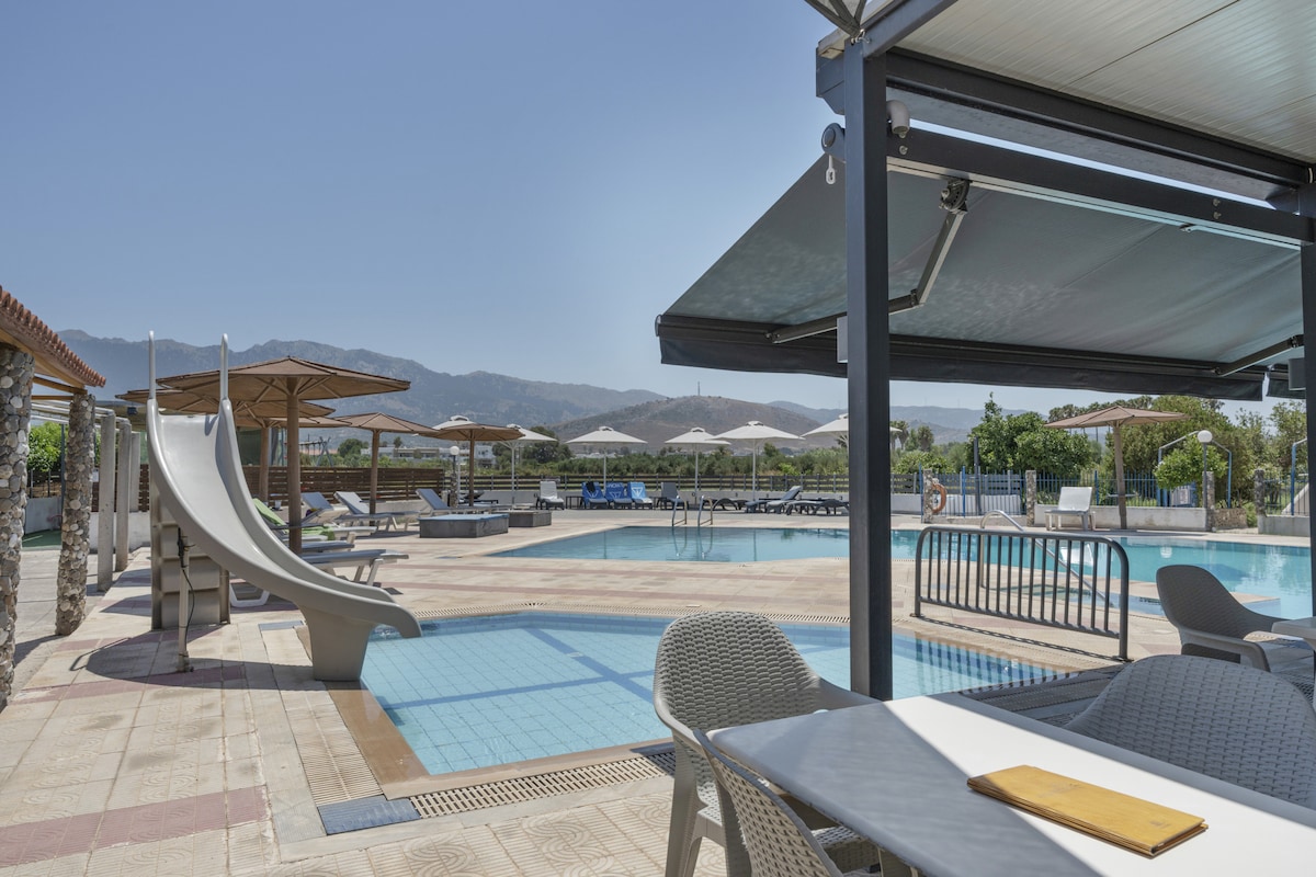 A shared outdoor pool area is visible with a water slide, lounge chairs, and umbrellas positioned around the pool. Tables and chairs are situated nearby, providing space for relaxation. The backdrop features distant mountains under a clear blue sky.