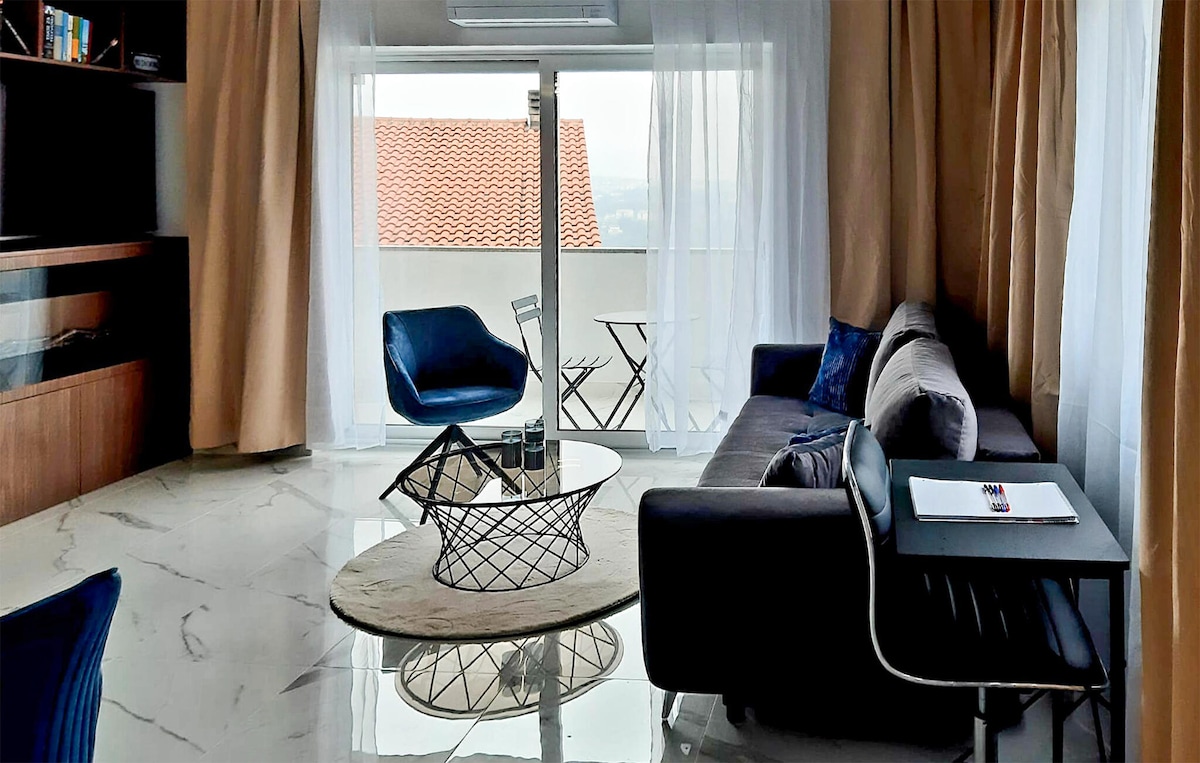 The living area features a modern grey sofa and a stylish black table, complemented by cozy blue chairs. A round rug adds softness to the space, while large windows with sheer curtains allow natural light to fill the room. A small outdoor table and chair set is visible on the balcony.