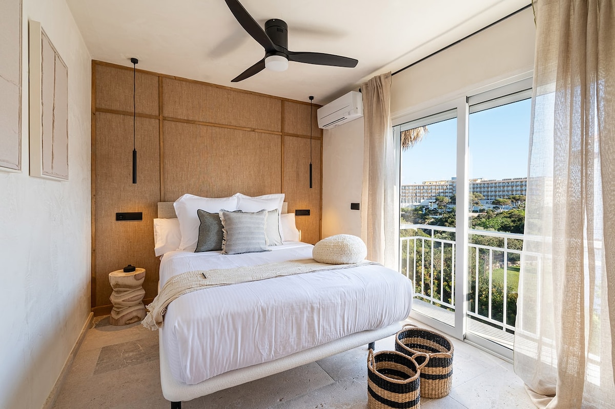 A bright bedroom features a spacious bed adorned with neutral-toned linens and decorative pillows. Natural light enters through large sliding doors, accompanied by sheer curtains. A ceiling fan is mounted above, and woven baskets are placed beside the bed, adding texture to the serene space.
