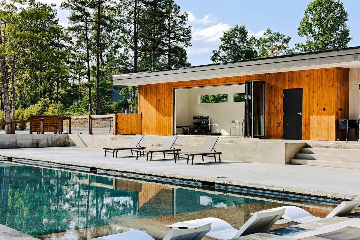 A modern building with wooden accents and large glass doors opens directly to a pool area. Lounge chairs are positioned beside the pool, reflecting the surrounding trees and sky. The space offers a serene ambiance for relaxation and outdoor gatherings.