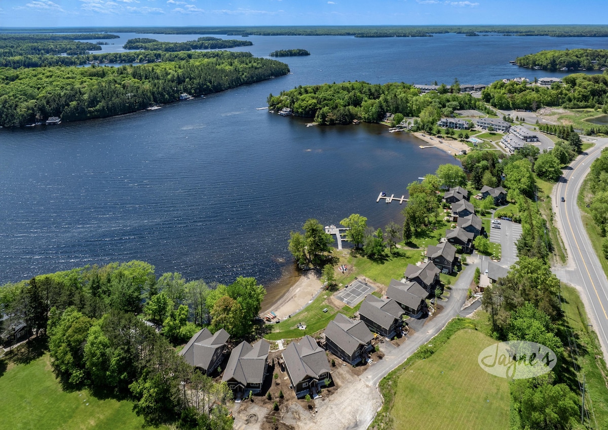 Aerial view of the resort showcasing multiple duplex villas lined along the shoreline. Sandy beach and shared docking areas are visible, along with the expansive waters of Lake Muskoka and surrounding greenery. The peaceful landscape emphasizes proximity to nature and outdoor activities.