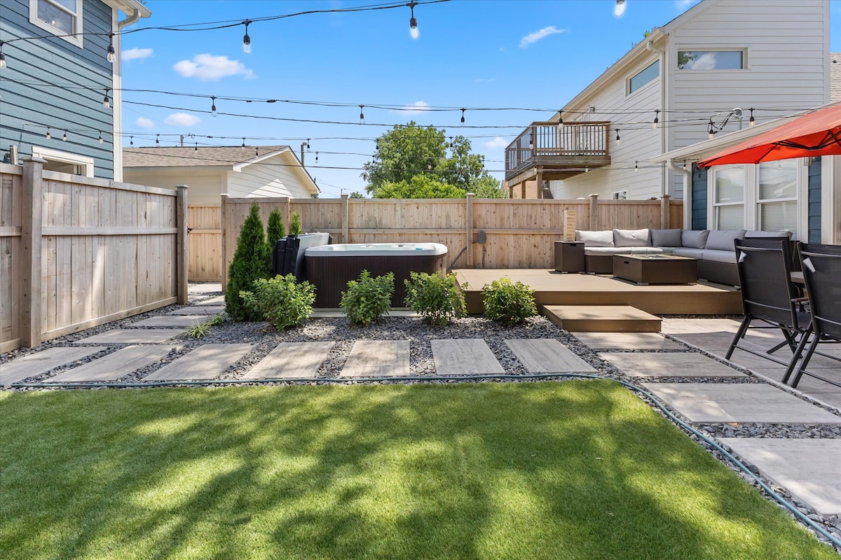 A fully fenced backyard features a hot tub surrounded by landscaped greenery and paved pathways. A wooden deck with seating is present, accompanied by an umbrella-shaded area. String lights above the seating area create a relaxed outdoor ambiance, complementing the well-maintained lawn.