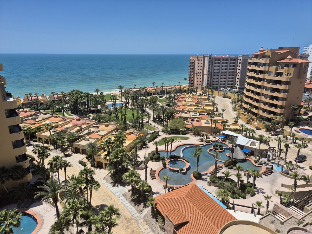 A panoramic view of the tropical landscape is visible, featuring lush palm trees and a spacious resort area. Multiple swimming pools are interspersed among the palm trees, and the ocean can be seen in the background, complementing the vibrant colors of the surroundings.