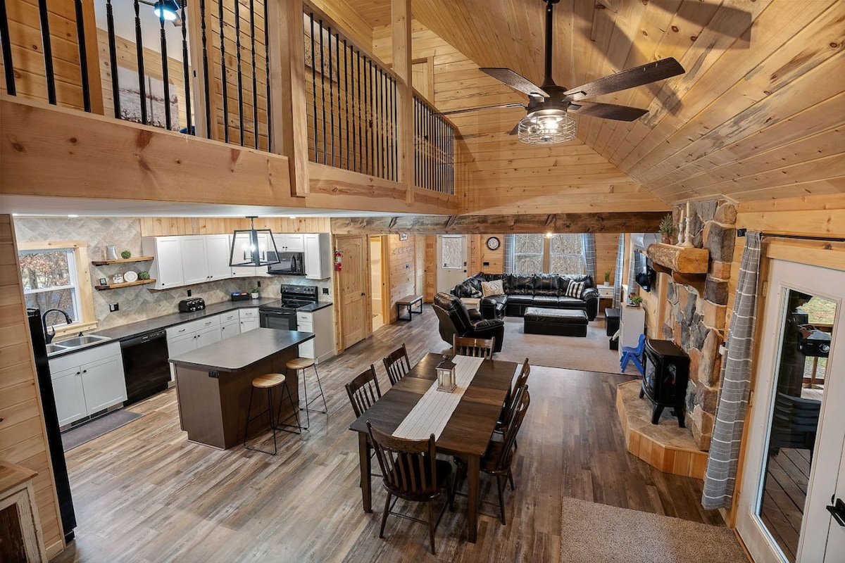 An open-concept living area is showcased, featuring high wood-paneled ceilings and a large kitchen with modern appliances. A spacious dining table accommodates multiple guests, while a comfortable leather couch is positioned around a stone fireplace, creating a cozy gathering space filled with natural light.