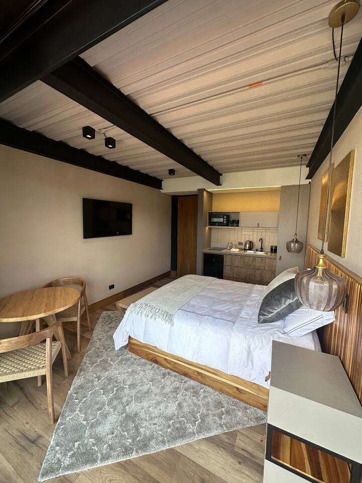A modern loft space features a spacious bed with neutral bedding, complemented by a decorative throw. A dining area with a round table and two chairs is present. The kitchenette is equipped, and a wall-mounted TV adds convenience. Light-colored walls and wood accents create a cohesive aesthetic.