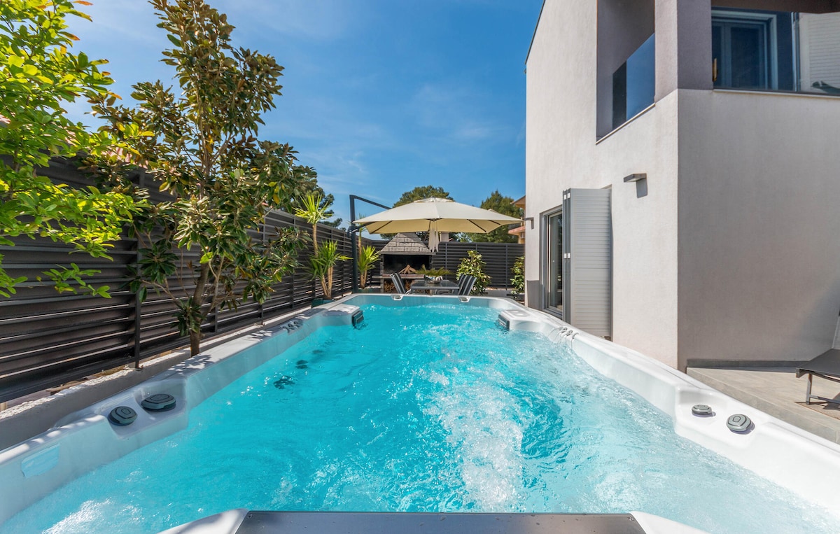 A heated outdoor pool is featured, inviting relaxation with counter-current swimming options. Sunlight reflects on the clear, turquoise water, surrounded by lush greenery. A sun umbrella and poolside chairs are positioned nearby for leisure and comfort.