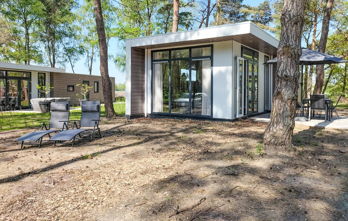 The vacation home features a modern design with large glass doors, allowing natural light to flow in. Two sun loungers are positioned in front, providing a space to relax in the outdoors. Tall trees surround the property, creating a tranquil setting.