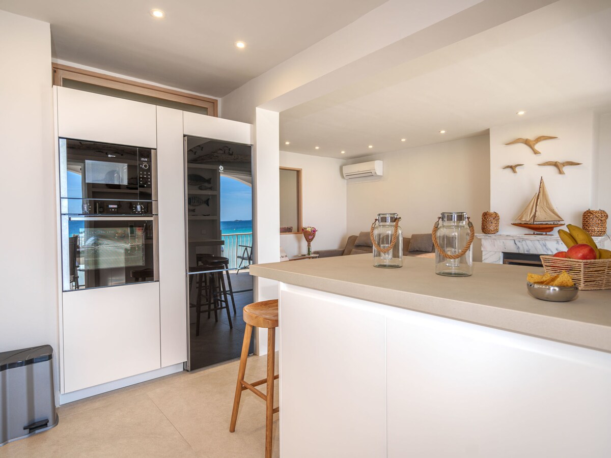 A bright kitchen area features modern appliances integrated into sleek cabinetry. A bar counter with a high stool is positioned nearby, offering a casual dining space. Natural light illuminates the room, while the scenic view of the sea is visible through the adjacent glass door.