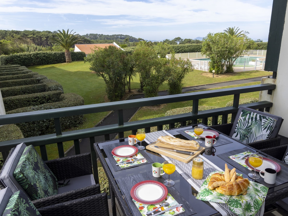 A balcony table is set for breakfast with plates, cups, and glasses, featuring croissants and orange juice. The view includes a neatly maintained garden and a swimming pool beyond. Surrounding greenery and palm trees create a serene atmosphere.