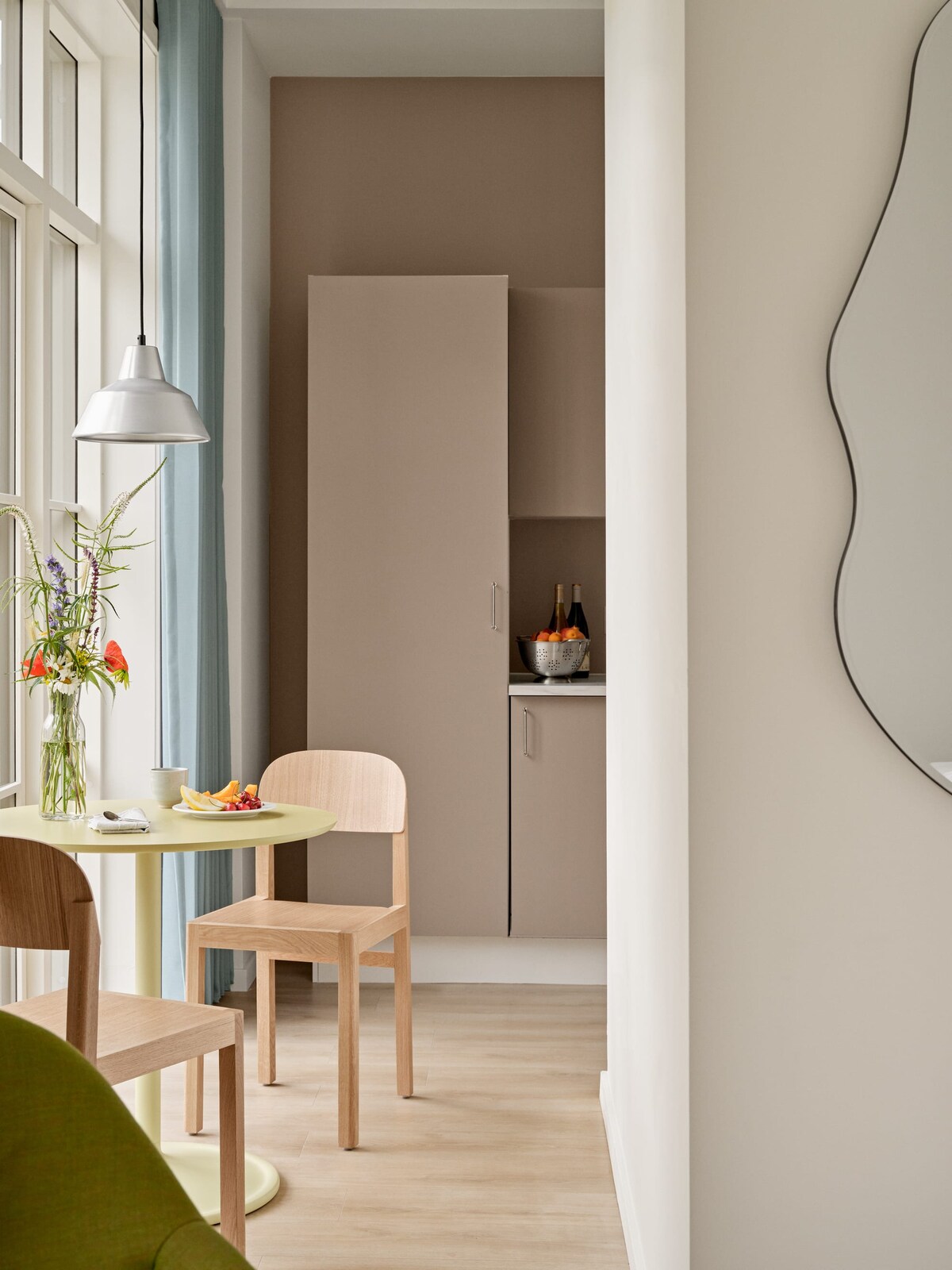 A cozy dining area features a round table topped with a small breakfast arrangement, complemented by two wooden chairs. The kitchen cabinetry in soft muted tones is seamlessly integrated into the space, while natural light filters through large windows draped in light curtains.