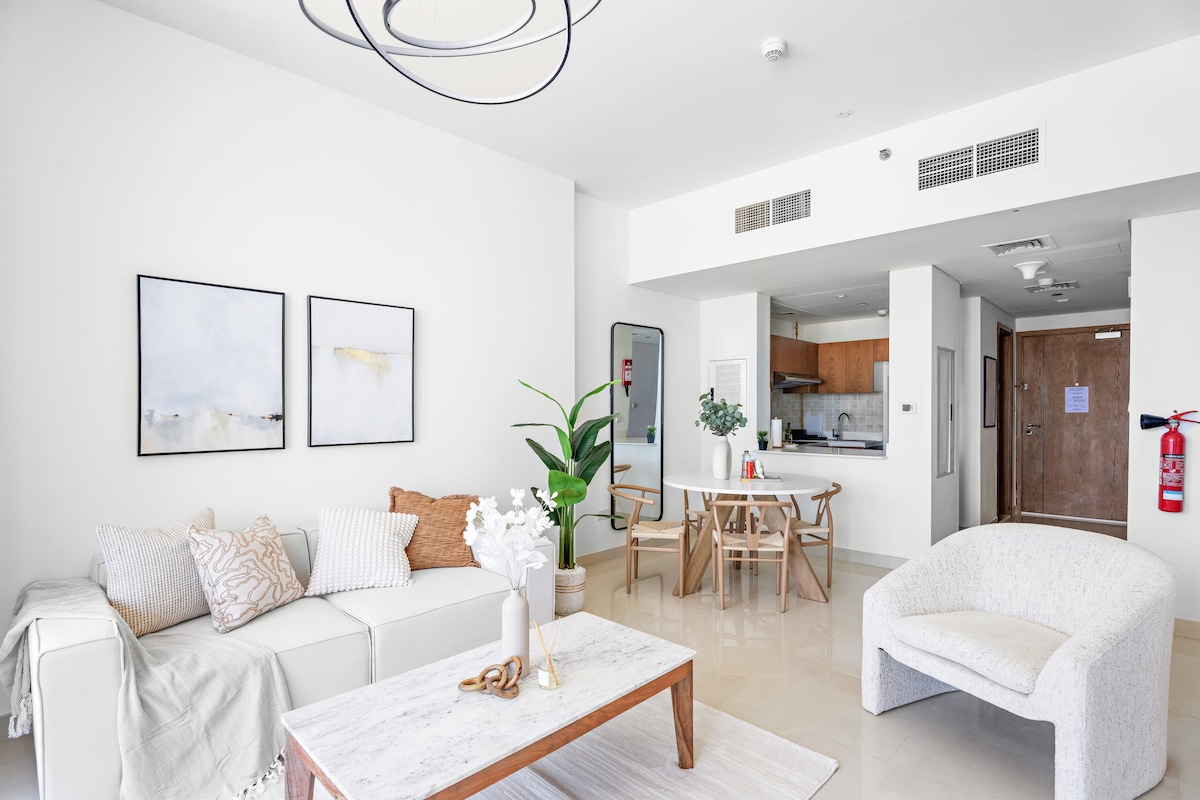 A modern living room is showcased, featuring a white sofa adorned with decorative pillows. A marble coffee table sits centrally, complemented by a minimalist dining area with wooden chairs. A large plant adds greenery, while abstract wall art enhances the space. Natural light fills the room.