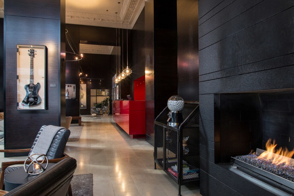 The lobby features sleek black walls complemented by elegant lighting and a cozy fireplace. A framed guitar artwork adds a musical touch, while a striking red reception desk stands out against the monochrome palette. Comfortable seating arrangements invite relaxation.
