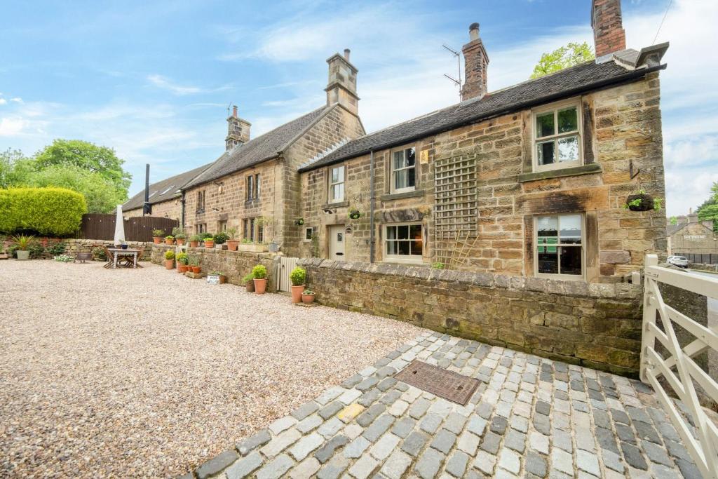 The exterior of Well Farm Cottage is showcased, featuring traditional stonework and charming windows. A gravel area in the foreground includes potted plants, while the lush green landscape is visible in the background, creating a serene rural setting.
