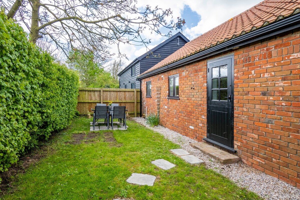 A private garden area is visible, featuring a well-manicured lawn with a stone pathway leading to a set of outdoor furniture that seats four. The garden is bordered by greenery, providing privacy and a calm environment. The exterior of the barn conversion is constructed from red brick.