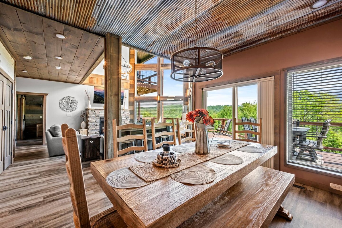 A spacious dining area features a large, rustic wooden table surrounded by chairs. Natural light filters through large windows, revealing views of the outdoors. Decor accents include a vase of flowers and woven placemats on the table, enhancing a welcoming ambiance.