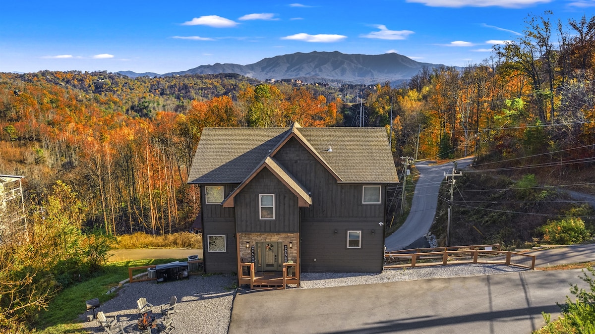 A newly built cabin sits against a vibrant autumn landscape, framed by colorful foliage and distant mountains. The structure features a rustic exterior with a combination of dark wood and large windows, while a spacious gravel driveway leads to the entrance.