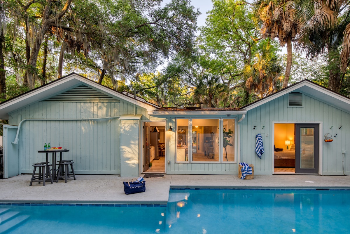 A modern home with light blue siding is framed by lush trees, with a private pool in the foreground. Glass doors provide a view into the living area, which is softly illuminated. An outdoor seating area with two chairs is positioned near the pool.