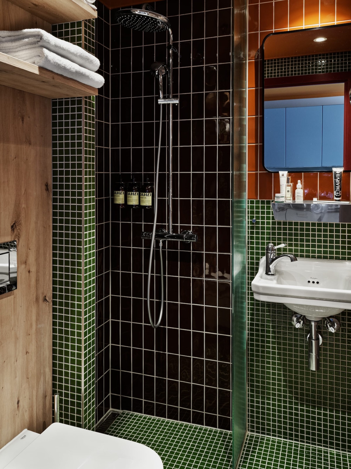 A modern bathroom design is highlighted by vibrant tiled walls in shades of green and brown. A glass shower enclosure is present, featuring a sleek rainfall showerhead. A compact sink is mounted on the wall, with eco-friendly toiletries arranged neatly beside a small mirror.
