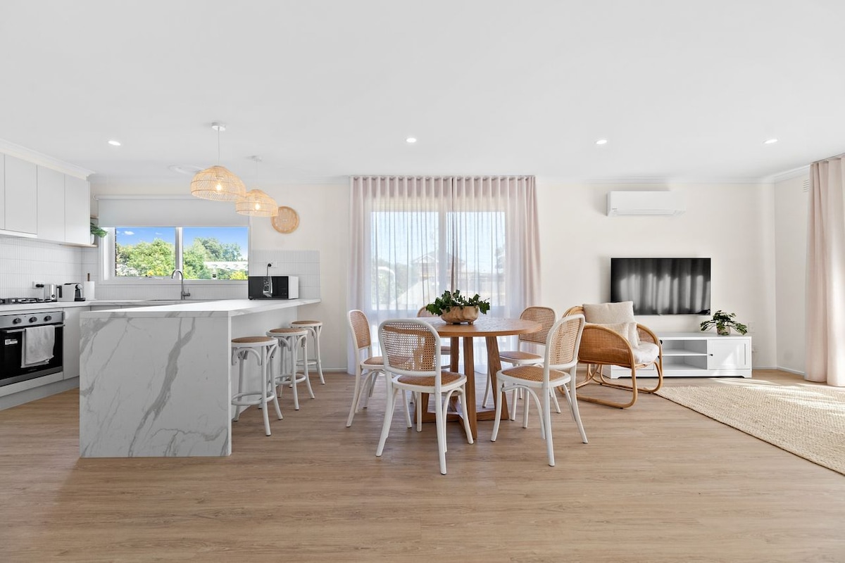A contemporary kitchen and dining area features light wooden flooring and is illuminated by natural light. A round dining table with white chairs is set near large windows, while the kitchen showcases marble accents, modern appliances, and wicker light fixtures overhead.