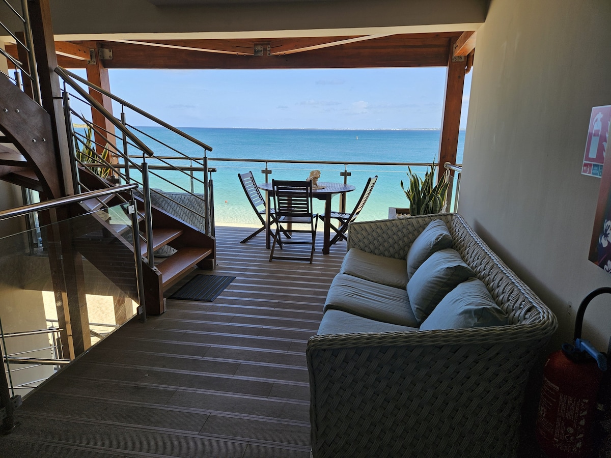 A private terrace features a light-colored woven sofa and a wooden dining table surrounded by four chairs. The sea view is unobstructed, showcasing clear blue water extending to the horizon, with gentle waves lapping at the shore.