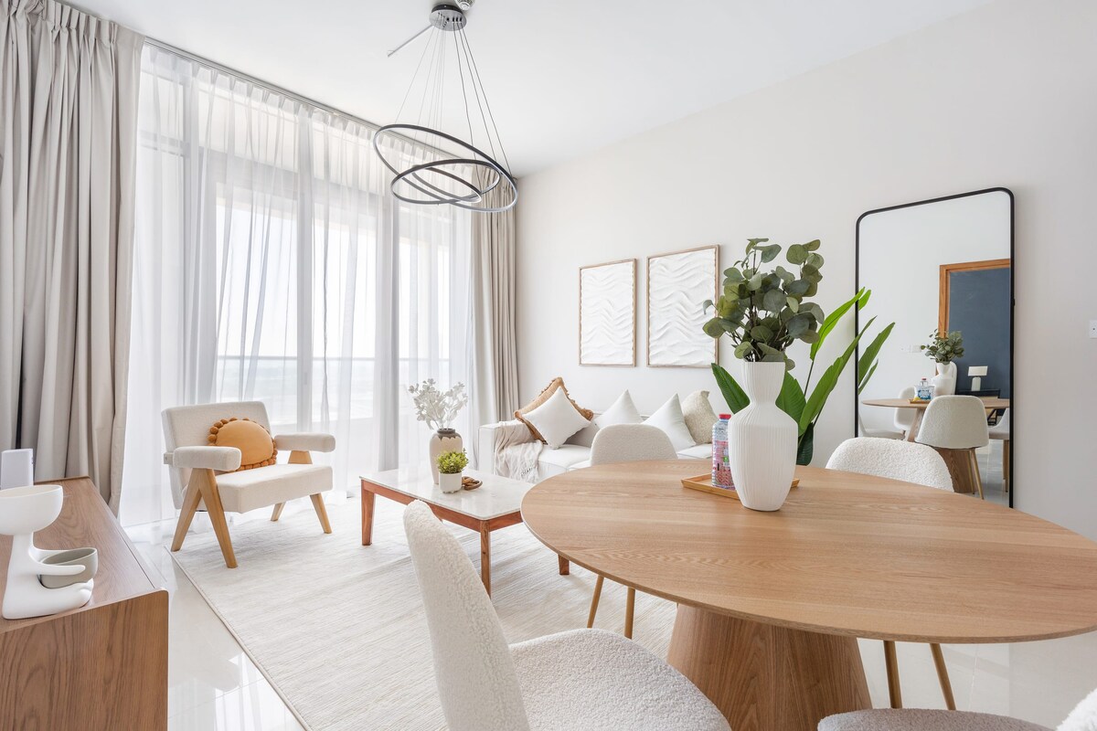 A bright living area features a circular dining table surrounded by upholstered chairs. A cozy seating arrangement is situated nearby, accented by a stylish mirror and wall art. Sheer curtains allow natural light to fill the space, enhancing the inviting ambiance of the room.