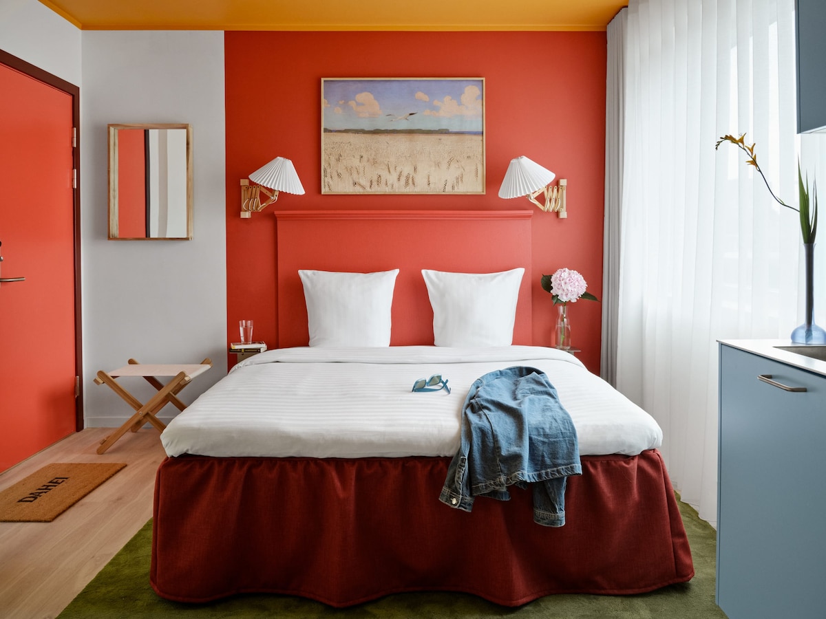 A bright and colorful studio features a spacious bed adorned with crisp white linens and two fluffy pillows. The walls are painted a warm red, contrasted by a vibrant yellow ceiling. A decorative artwork hangs above the bed, complementing the welcoming atmosphere.