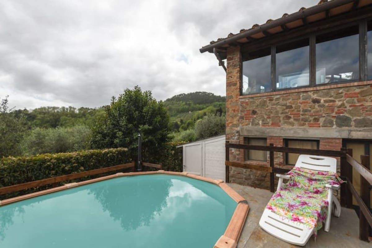 A private pool is positioned beside a rustic stone building, surrounded by lush greenery. A lounge chair is nearby, adorned with a colorful towel, while the scenic view includes rolling hills in the background under a partly cloudy sky.