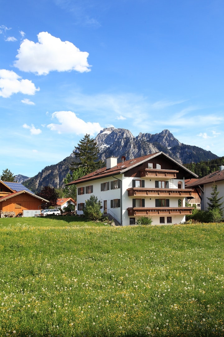 A multi-story building is situated amidst a lush green meadow, surrounded by residential homes. The backdrop features a prominent mountain range under a clear blue sky, with scattered clouds enhancing the natural scenery.