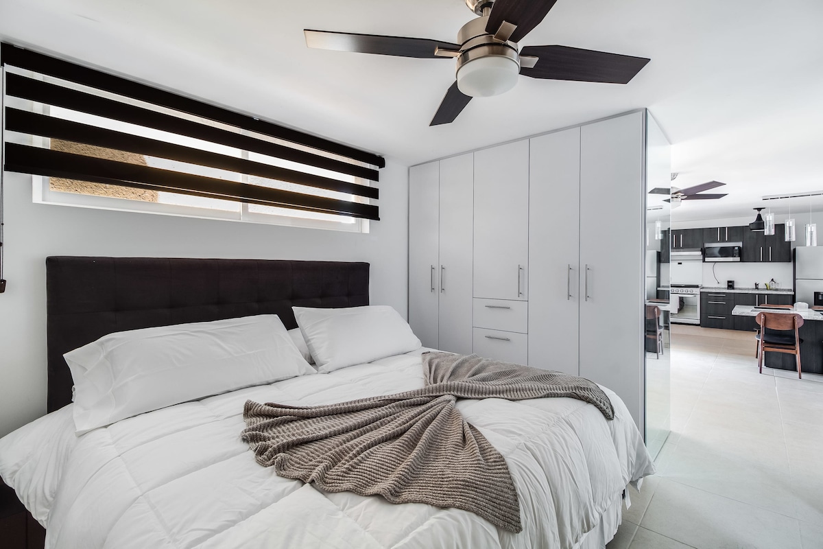 A spacious bedroom features a large bed draped with a soft blanket, complemented by modern bedding. Vertical blinds filter natural light, while a ceiling fan circulates air. A sleek closet offers ample storage, and an open-concept kitchen and dining area are visible in the background.