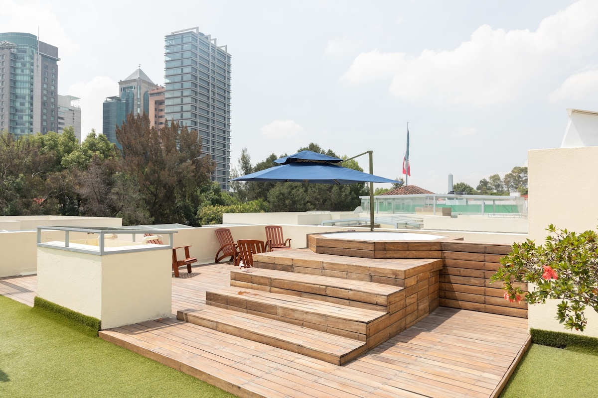 A spacious rooftop terrace features wooden decking with steps leading to a raised platform. Comfortable wooden chairs are arranged under a large blue umbrella, providing shade. Surrounding greenery enhances the urban view of nearby buildings.