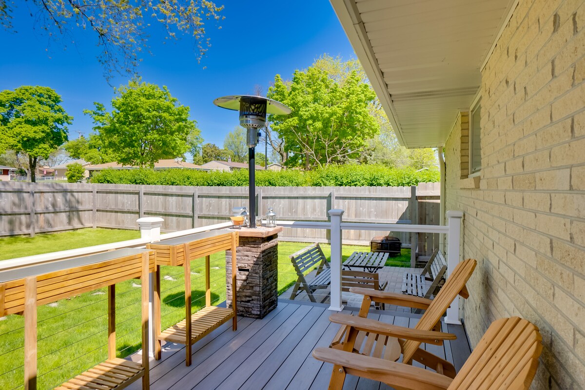 An outdoor deck offers comfortable seating with wooden chairs and a dining table. A patio heater stands near the table, surrounded by a green lawn and well-maintained hedges. The space is enclosed by a wooden fence, creating a private atmosphere.