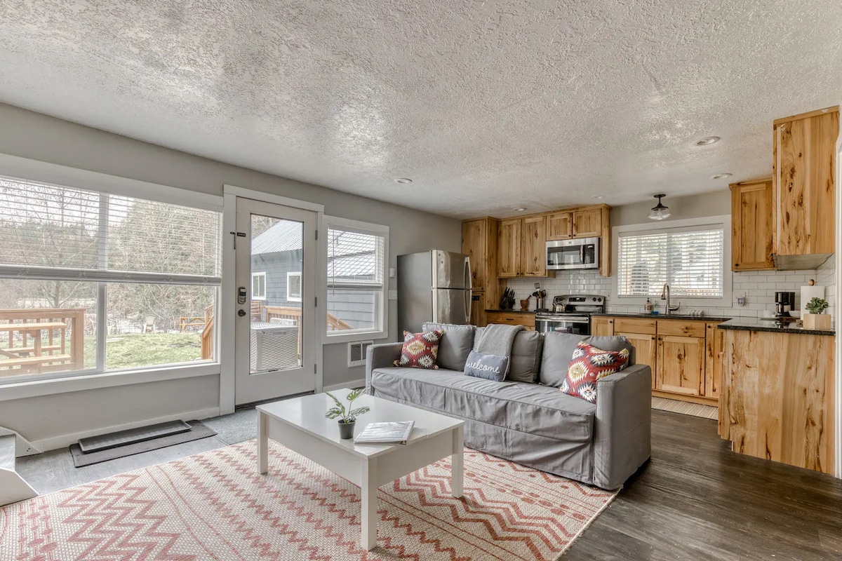 A spacious lounge area showcases a comfortable grey sofa adorned with decorative throw pillows. Natural light floods in through large windows, illuminating the modern kitchen with stainless steel appliances. A dining table is visible, and a textured area rug adds warmth to the open space.