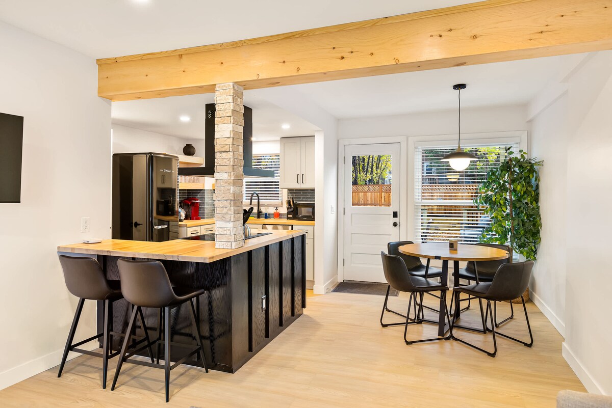 The spacious kitchen features modern cabinetry, stainless steel appliances, and a large island with seating for three. A dining table for four is positioned near large windows, allowing natural light to illuminate the area. A potted plant adds a touch of greenery.