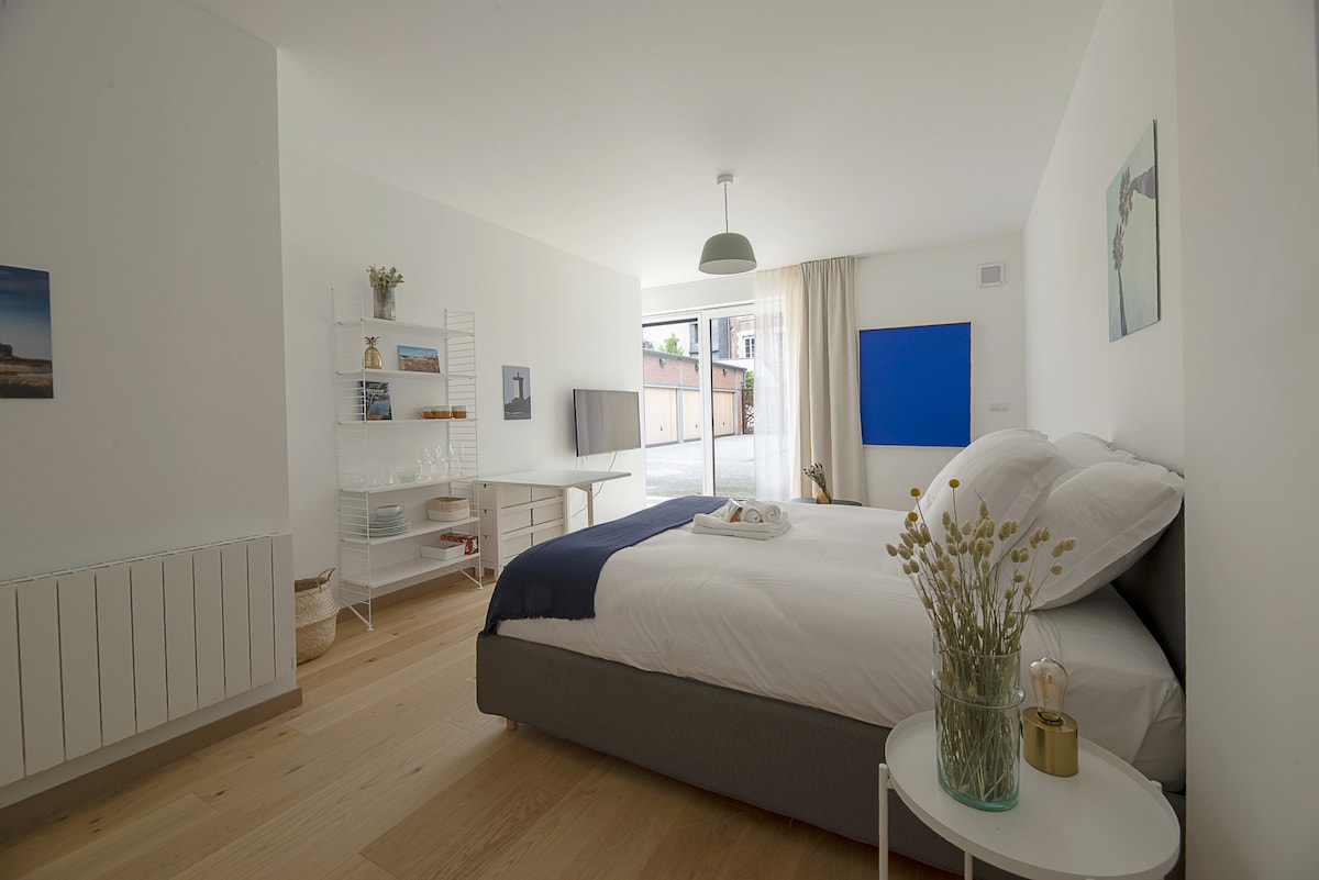 A spacious studio is highlighted by a large queen size bed topped with neutral linens. Natural light fills the area through a glass door leading to the courtyard. A minimalist shelving unit stands against the wall, complemented by simple decor and a blue accent wall.