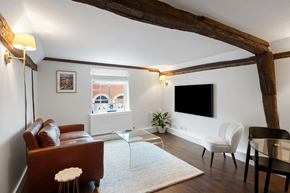 The living area features historic wooden beams and a large window with natural light. A cozy leather sofa is positioned opposite a flat-screen TV mounted on the wall. A glass coffee table rests on a soft area rug, complemented by a simple chair and a dining table.