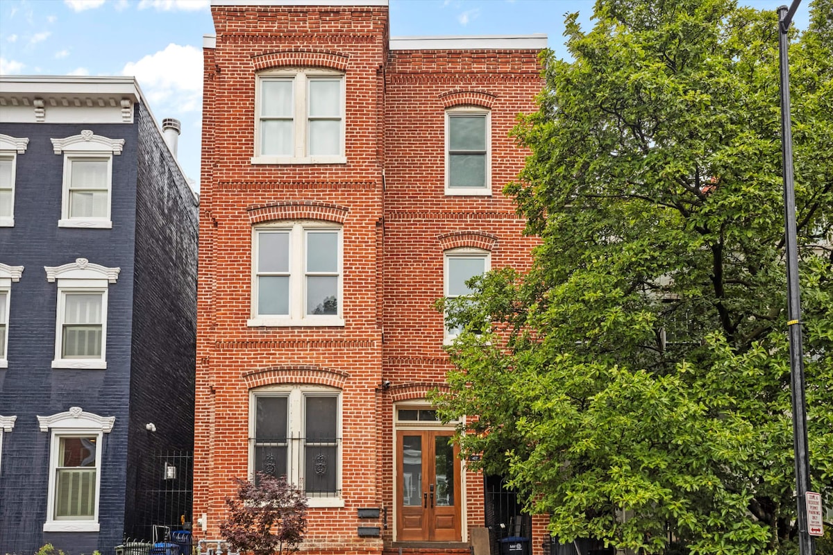 A historic brick townhome built in 1896 is surrounded by lush greenery. Large windows reflect ample natural light, and a welcoming entrance is framed by well-maintained landscaping. The building showcases traditional architectural details, blending seamlessly with the vibrant neighborhood.