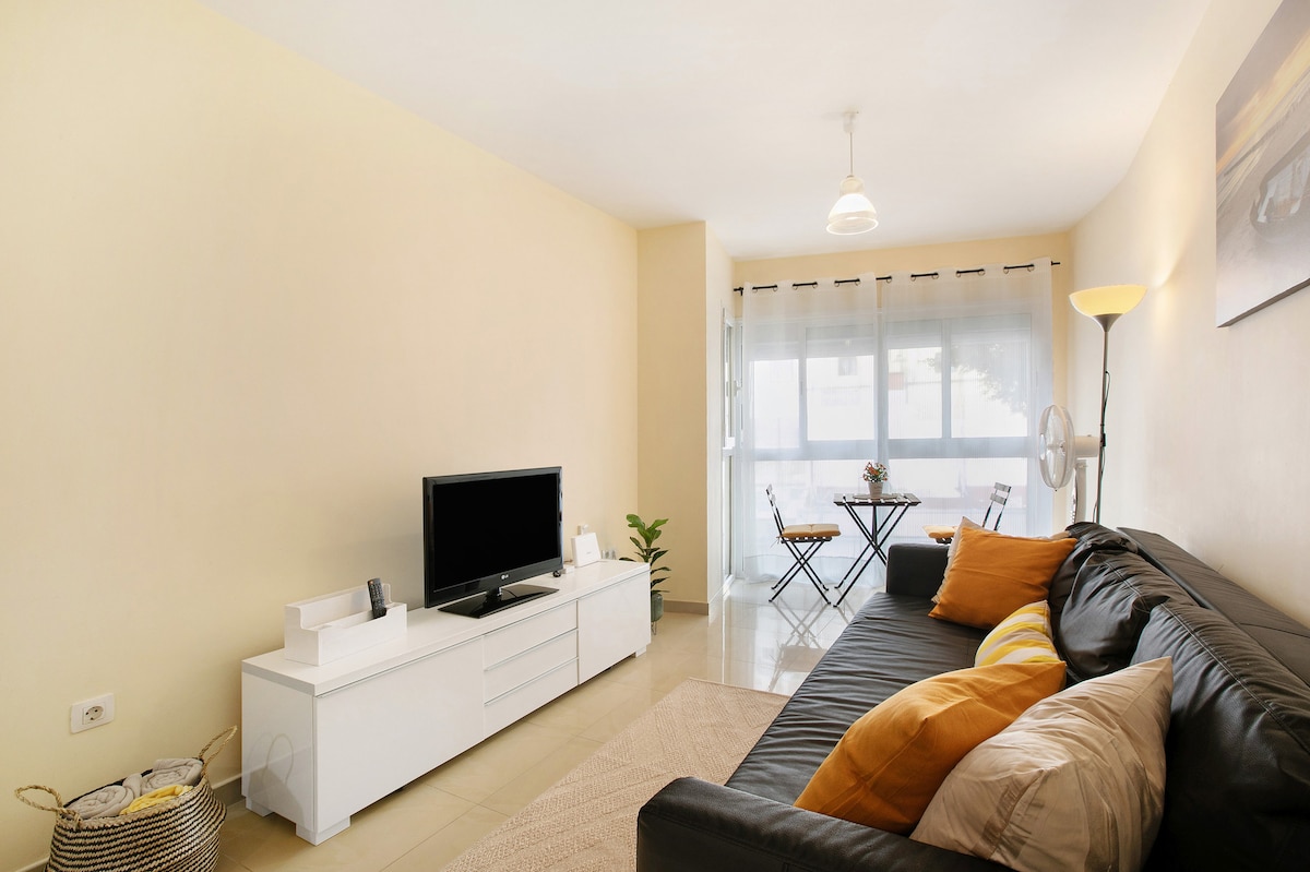 The living room features a modern design with a black sofa adorned with yellow cushions. A white TV stand holds a flat-screen television, while natural light filters through the large glass doors leading to a small dining area with two chairs and a table.